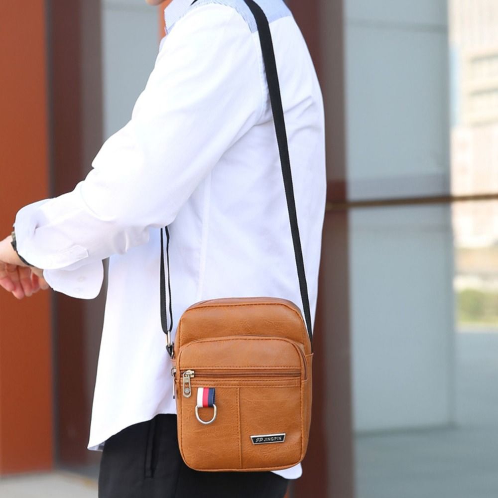 Hellbraune Ledertasche im Sling-Bag-Stil mit schwarzem Gurt und kleinem Flaggenemblem am vorderen Reißverschluss-Zipper, entworfen als Umhängetasche Schulter für den Freizeitgebrauch