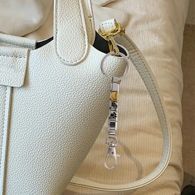 Llavero con dije de eslabones italianos con acabado plateado, con un dije de rosa en color dorado y detalles de huella de pata negros, sujeto a una correa de bolso blanco.