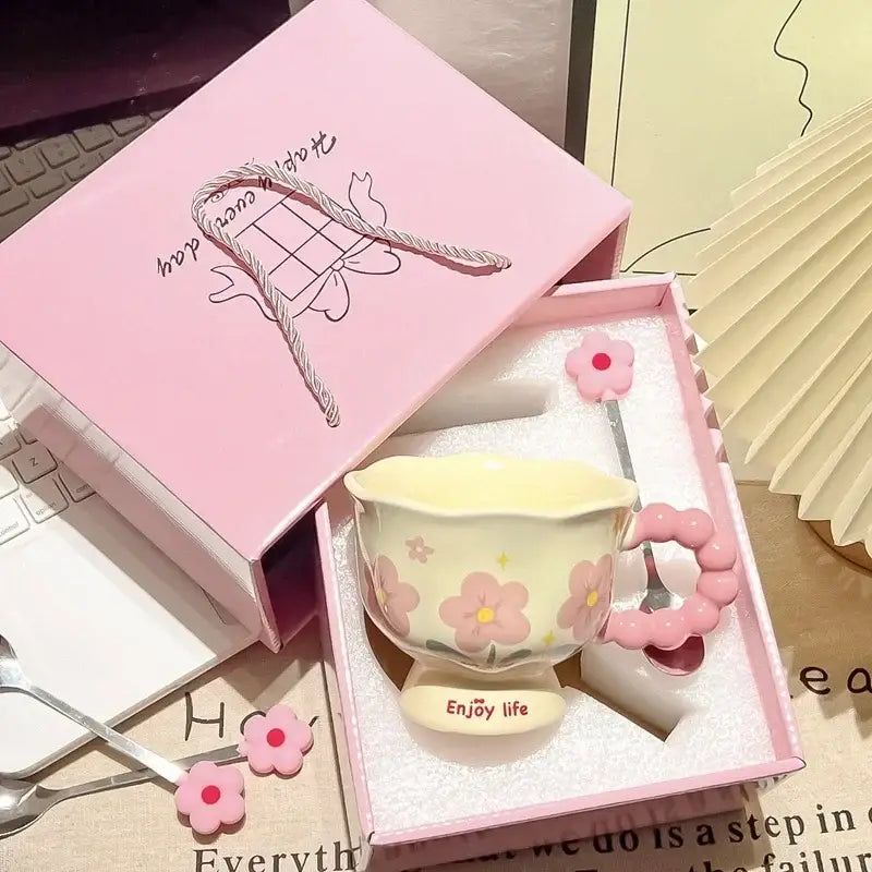 Une tasse tulipe en céramique jaune pastel avec des motifs floraux roses et une anse assortie en perles roses, placée dans une boîte cadeau rose.