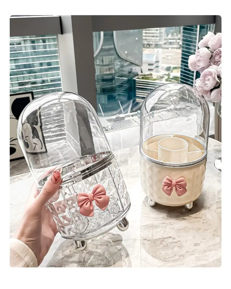 Boîte de rangement tournante pour maquillage et organisation des cosmétiques en forme de dôme en verre transparent, avec un accent de nœud rose et une bordure métallique argentée.