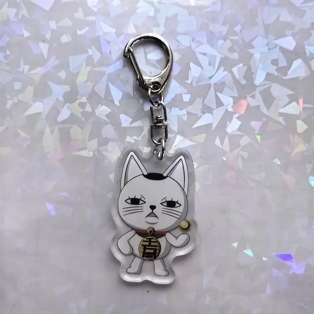 胸に「吉」と刻印された金色のコインを持つ、黒い目とヒゲの白いカートゥーン猫の、クリアアクリル『ダンダダン』アニメキーホルダー。