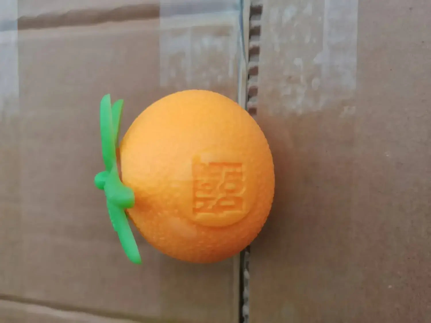 Eine Frontansicht eines orangefarbenen, runden quetschbaren Spielzeugs mit grünem Stiel und eingeprägtem Logo aus dem Erdbeere-Orange-Banane-Stressabbau-Set.