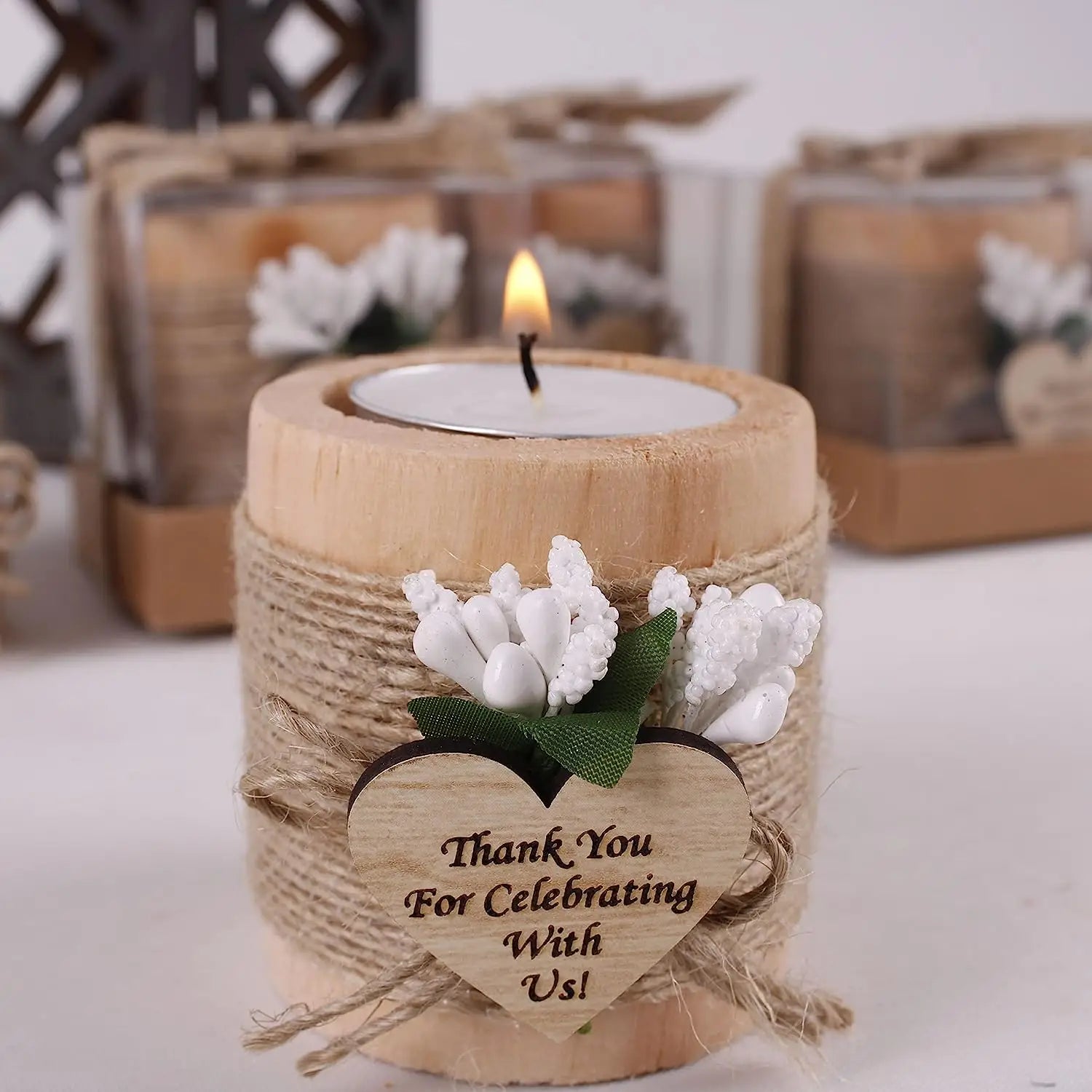 Un bougeoir rustique en bois pour petite bougie chauffe-plat, enveloppé de cordelette naturelle et orné de fleurs artificielles blanches, avec une étiquette en bois en forme de cœur portant le texte « merci de célébrer avec nous », utilisé comme cadeau pour les invités de mariage.