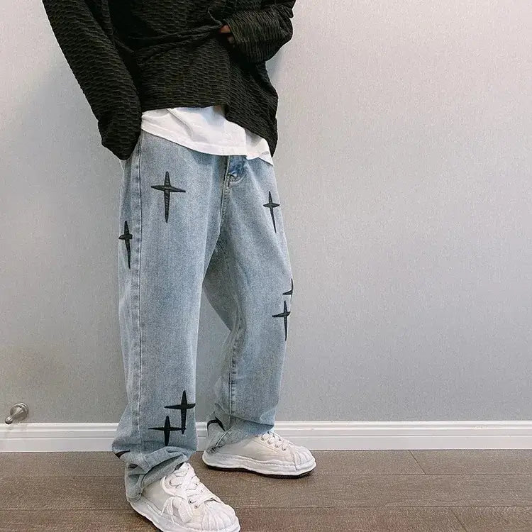 Jeans en denim délavé clair avec broderie croisée noire sur les jambes, présentant un design ample à jambes larges.