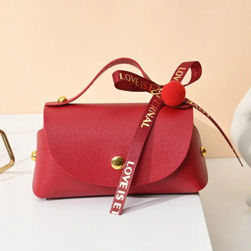 Mini sac à main en faux cuir rouge avec quincaillerie ton or et un nœud en ruban décoratif portant la mention « l’amour est éternel », conçu comme mini sac cadeau de mariage en cuir.