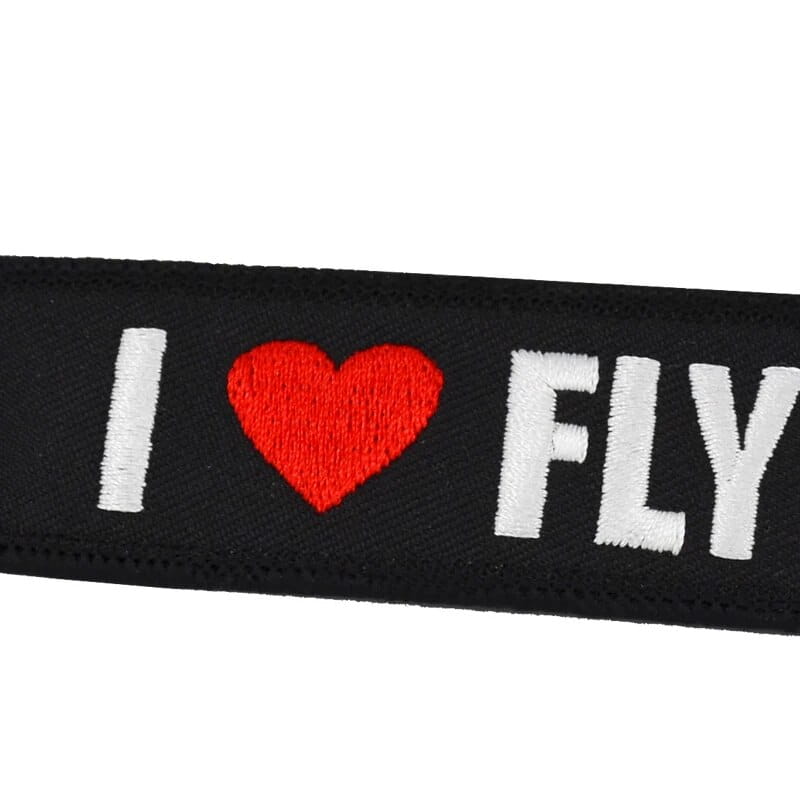 赤いハート記号を挟む形で、白いステッチ文字の「i」と「fly」が入った黒い布ベルトストラップを特徴とする「フライト前に取り外してください」刺繍キーチェーン。