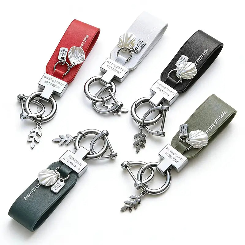 Juego de cinco llaveros de piel con funda en rojo, blanco, negro, verde oliva y gris oscuro, con herrajes metálicos de tono plateado, que incluye colgantes en forma de concha y dijes en forma de hoja para su uso como llavero antirrobo para automóvil.