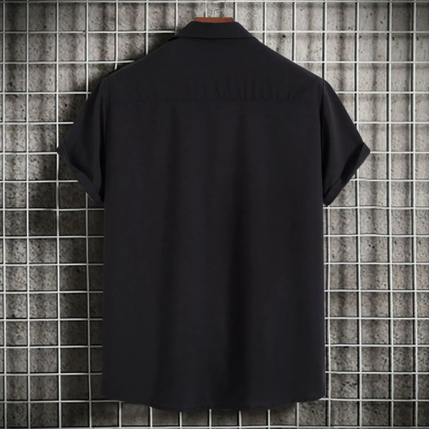 Chemise noire à manches courtes pour homme, avec col classique et design boutonné, suspendue sur un cintre en bois contre un mur à motifs quadrillés, convenant comme chemise de plage décontractée unie dans le style mode pour hommes.
