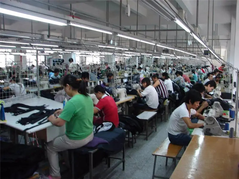 Reihen von industriellen Nähmaschinen mit metallischen Gestellen und weißen oder grauen Gehäusen, die entlang von Werkbänken in einer hell beleuchteten Fabrik angeordnet sind und Damen-Chiffonblusen zum Knöpfen mit floralem Muster herstellen