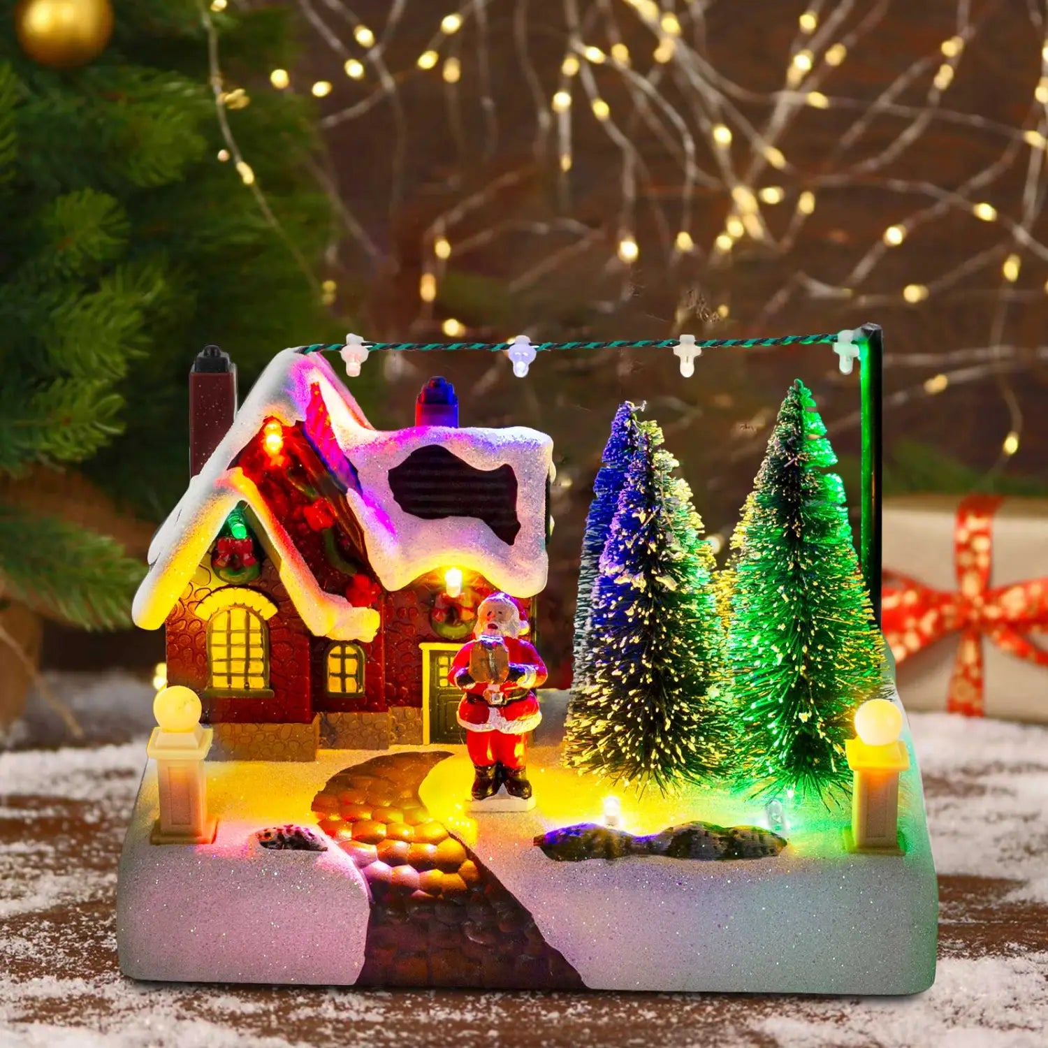 サンタと雪を備えたLEDクリスマスビレッジの家。雪の積もった屋根の赤い照明付きの家、光り輝く窓、色とりどりの松の木、そして雪の小道にサンタクロースの置物が配置された情景。