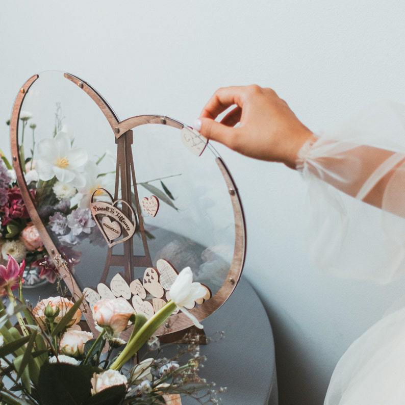 Boîte de dépôt pour livre d’or de mariage en bois en forme de cœur couleur or rose, avec des panneaux en acrylique transparent et des cœurs en bois suspendus à son cadre.