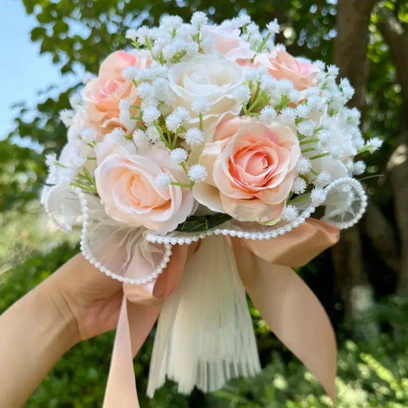 A delicate white silk rose wedding bouquet featuring peach and white artificial roses accented with babys breath and tied with a sheer lace ribbon adorned with pearl beads.