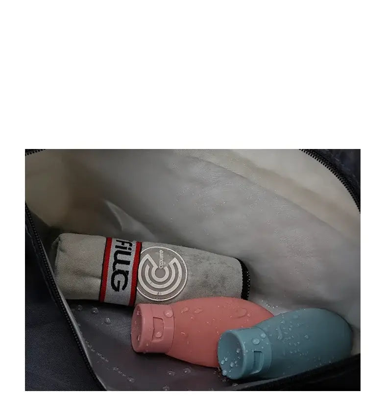 Un paraguas de tela gris enrollado con una banda a rayas roja y blanca y logotipo circular, junto con botellas de silicona de viaje rosa y teal, al lado de una bolsa de viaje tipo duffel acolchada tote weekender grande.