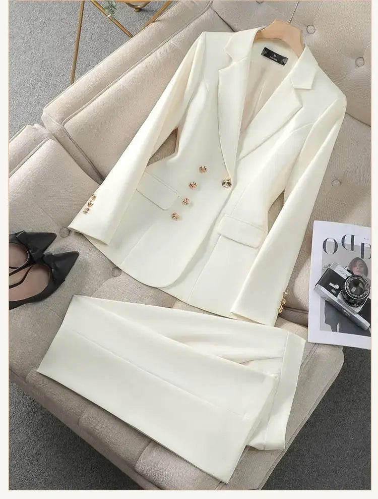 Ensemble de tailleur d’affaires pour femme cadre de couleur crème, composé d’une veste à boutons floraux ton-or et d’un pantalon assorti, présenté sur un canapé capitonné.