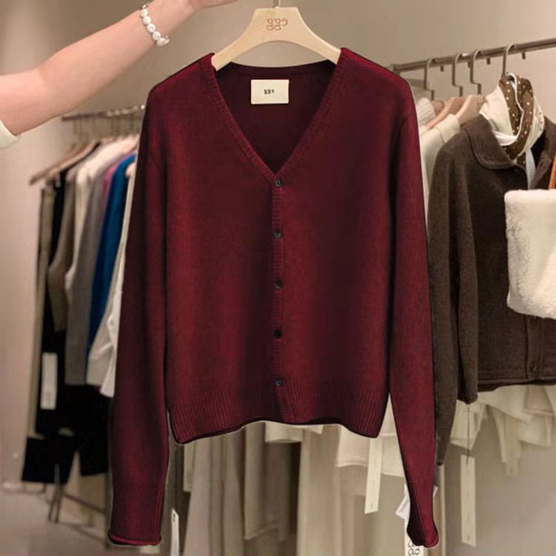 Un pull cardigan en laine mérinos bordeaux foncé à encolure en V, à manches longues et à boutons noirs, suspendu à un cintre en bois.