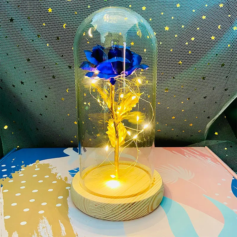 „Eternal Gold Rose Glass Dome LED Light“, mit einer leuchtend blauen Rose mit goldenem Stiel und Blättern, umschlossen in einer klaren Glaskuppel auf einem Holzsockel.