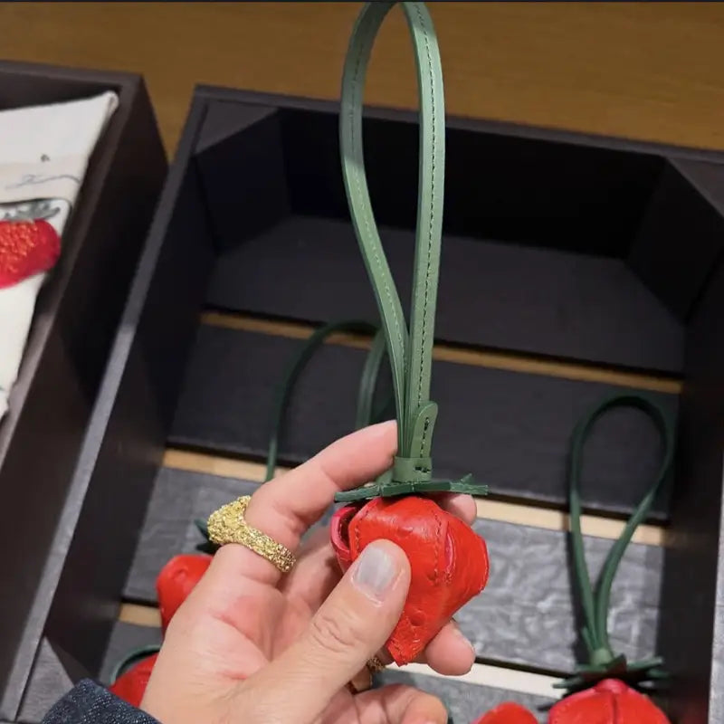 Un porte-clés en cuir en forme de fraise rouge, vibrant, avec une tige et une lanière vertes, présenté comme charm pour sac à main en cuir végétal et ornement de sac à main de créateur.