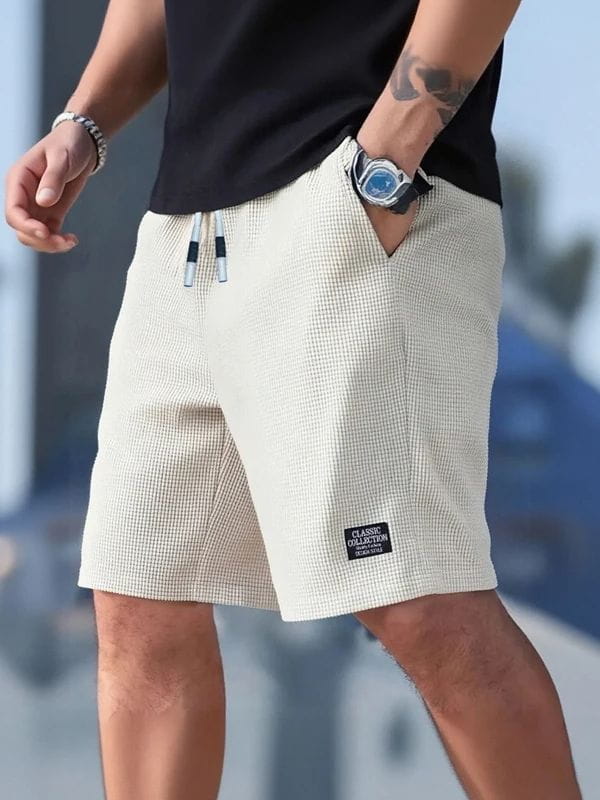 Herren-Waffelstrick-Freizeit-Badeshorts in Beige mit schwarzen und weißen Kordeln sowie einem kleinen Label „Classic Collection“ am unteren linken Bein.