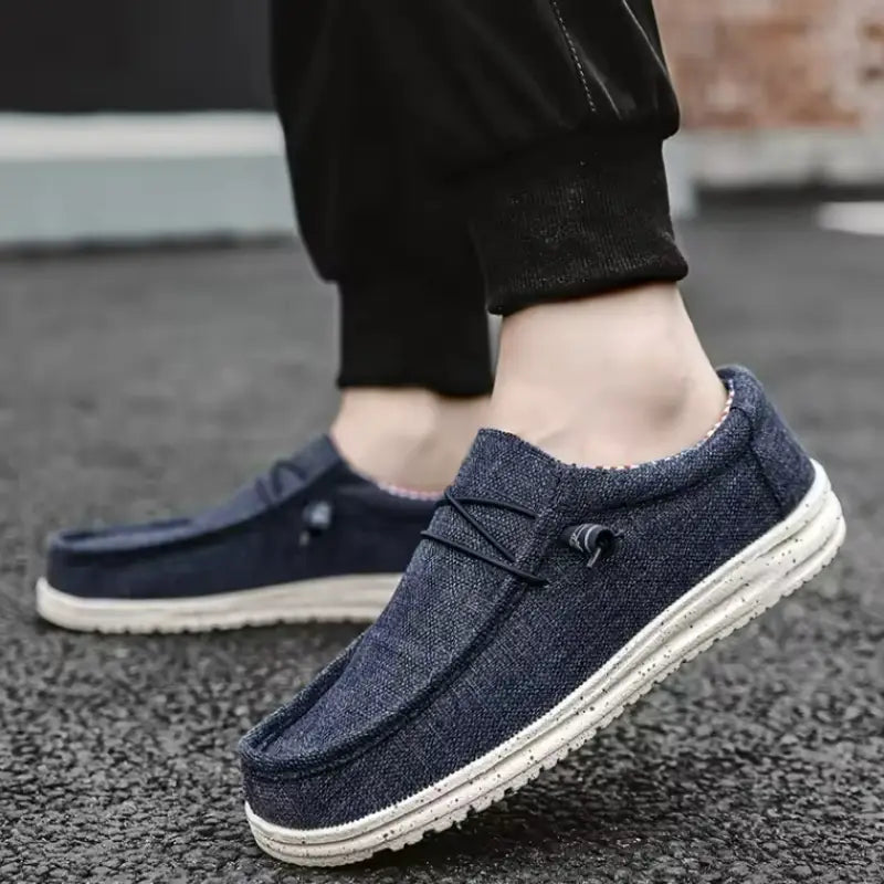 Chaussures décontractées de type mocassins à enfiler en toile pour hommes, de couleur bleu foncé, avec des semelles en caoutchouc blanches et des lacets noirs décoratifs.