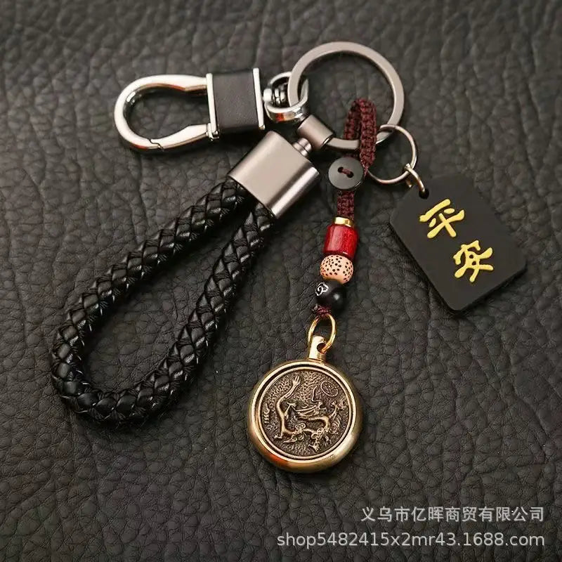 赤と黒のビーズでアクセントを施し、刻印された「龍」のデザインをあしらったブロンズ色の円形メダリオンと、小さな長方形のタグ（中国の干支をモチーフにした編みレザー製ラッキーペンダントのコレクション）付きの黒の編みレザー製キーチェーン。