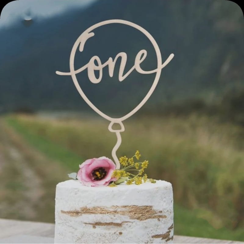 Un délicat topper de gâteau en bois clair, en forme de ballon, portant le mot « one » inscrit avec élégance à l’intérieur de son contour, pour une décoration florale d’anniversaire.