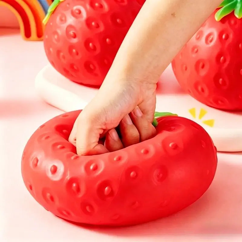A red strawberry-shaped stress ball with a textured surface and green leafy top designed as a jumbo slow rising sensory fidget toy.