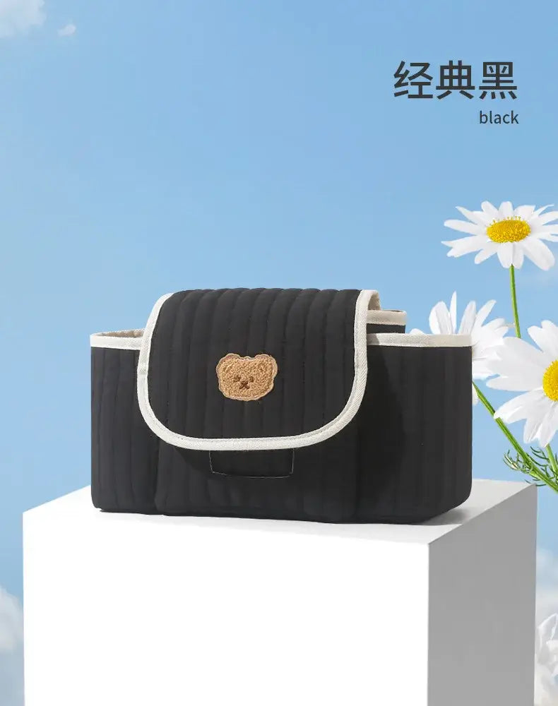 Schwarze gepolsterte Wickeltasche für Babys mit beigem besticktem Bären-Gesicht und weißem Besatz, gestaltet als Organizer-Beutel für den Kinderwagen.