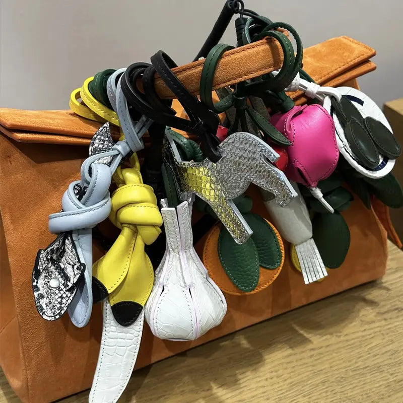 Un ensemble de charms d’animaux et de fruits en cuir végétal, fabriqués à la main et aux couleurs variées, suspendus à une poignée de sac en daim camel, conçus comme ornements de sac à main de créateur.