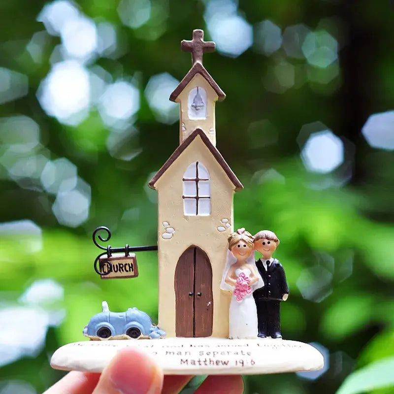 Figura decorativa de boda en resina color beige con iglesia, techo y puertas color marrón, que presenta a una novia con vestido blanco sosteniendo un ramo de flores rosadas junto a un novio con traje negro; incluye un coche azul pequeño sobre la base, ideal como regalo de aniversario.