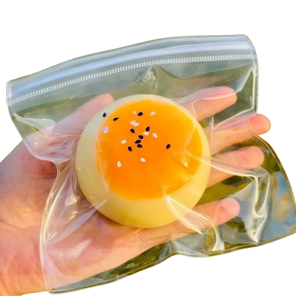 Ein durchscheinendes, rundes Silikon-Eigelb-Gebäck, langsam aufsteigend, als knetbares Spielzeug, mit einem kräftigen orangefarbenen Eigelb und einem Belag aus weißen Sesamsamen, von vorn betrachtet.