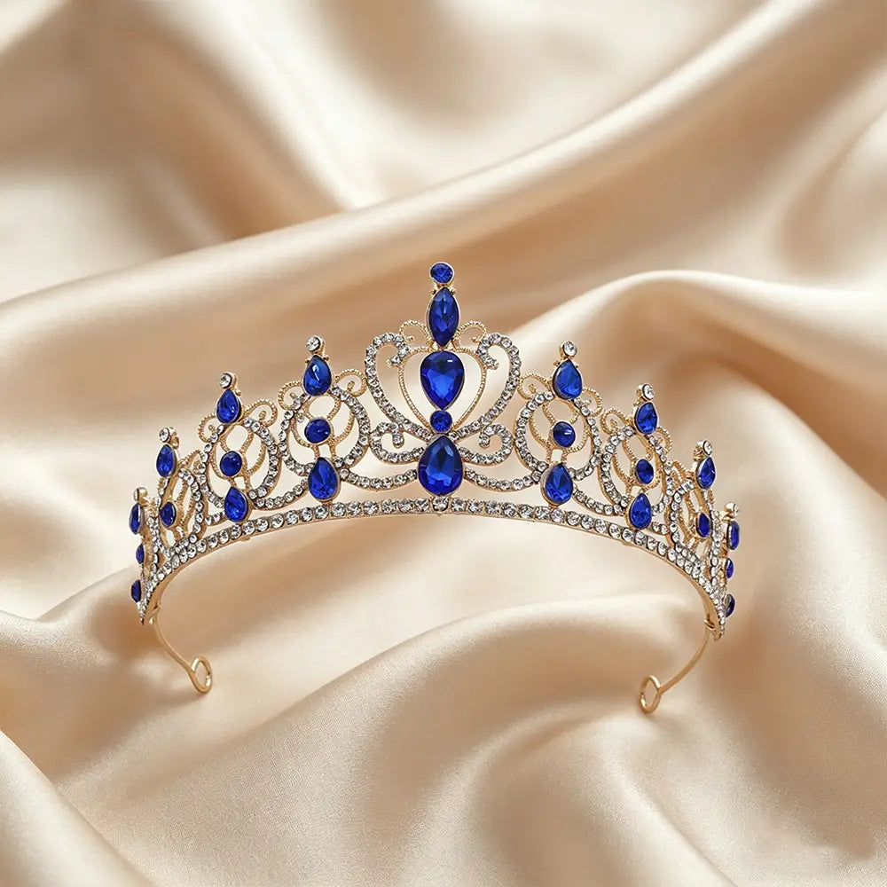 Un accessoire de coiffure de mariée en couronne de diadème baroque en strass doré, orné de cristaux profonds bleus en forme de goutte et de petits strass transparents, sur un fond de satin beige doux.