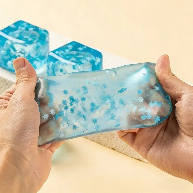 ein blauer durchscheinender Eiswürfel mit Schneeflocken-Motiv zur Stressreduktion aus Harz, mit eingebetteten schneeflockenförmigen Partikeln, in den Händen gehalten.