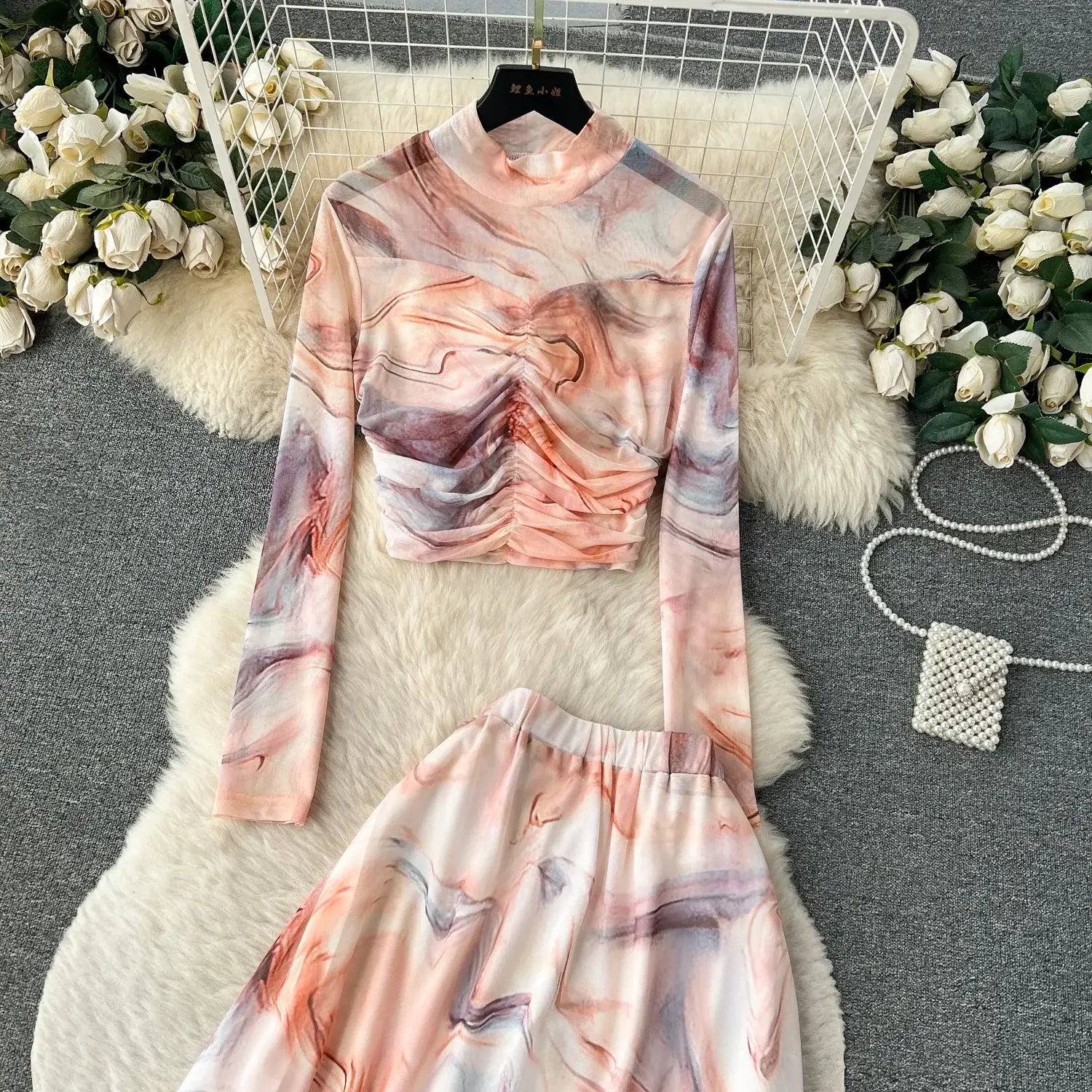 A soft long-sleeved high-neck top with a marbled pink and gray pattern hangs on a black hanger featuring an elastic closure type as part of the two piece mesh skirt set marble print.