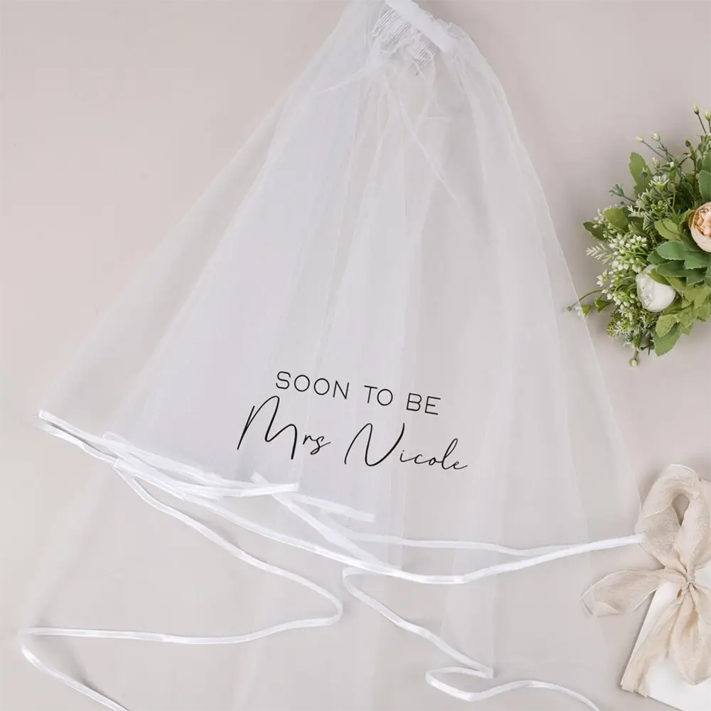 繊細な白いチュール製のパーソナライズされたバチェロレッテパーティー用ベールで、「soon to be mrs nicole」という刻印がエレガントな黒の筆記体で入り、縁に沿って細いサテンのリボンの結び紐が施されています。