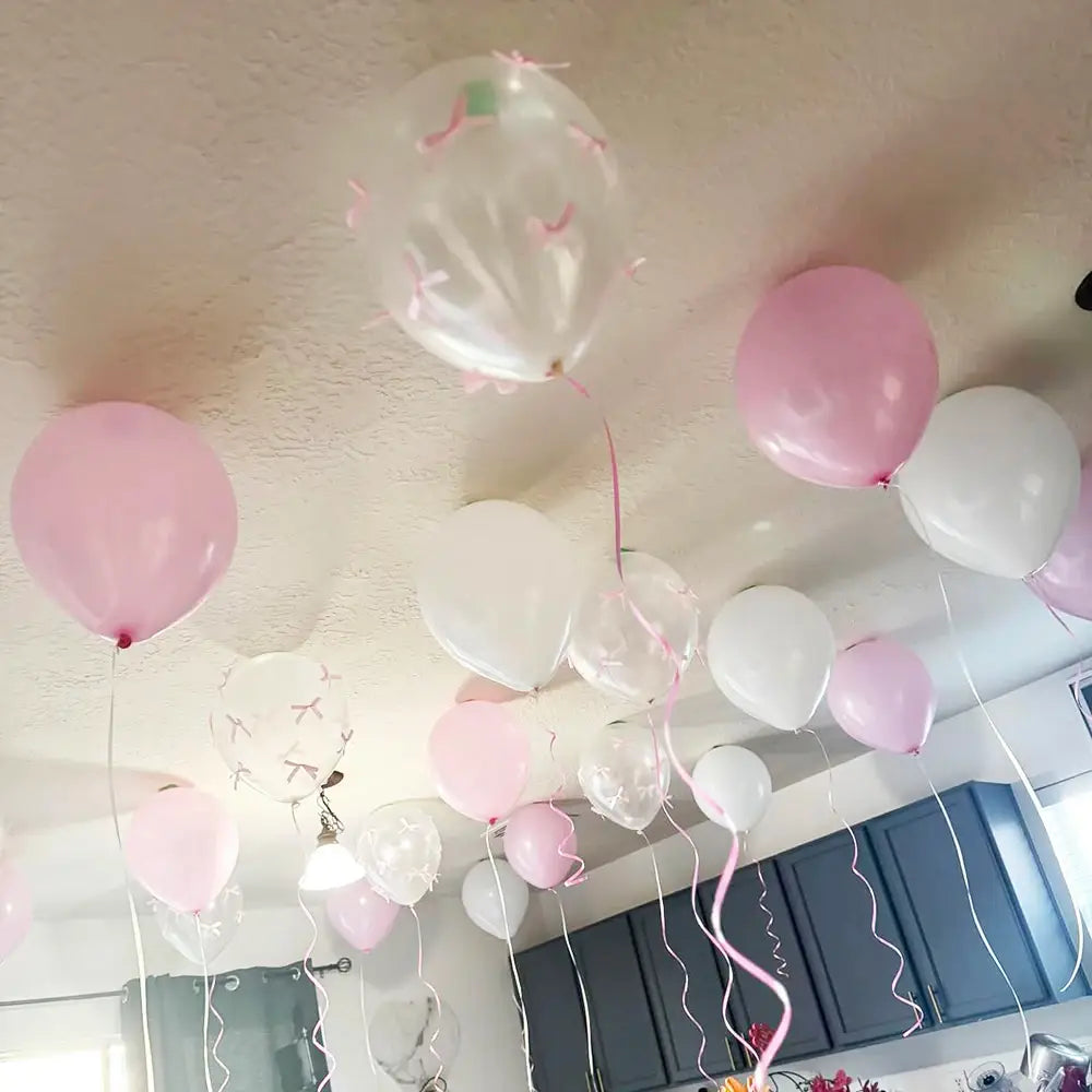 Un ensemble de ballons à nœud blanc avec des rubans roses, fabriqués en latex, flottant près du plafond, pour la décoration de fête.