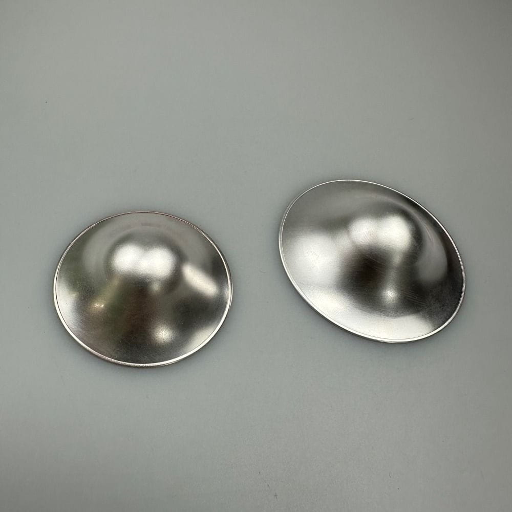 Ein Paar glänzender, metallisch-silberner Brustwarzen-Schutzvorrichtungen mit glatten, reflektierenden Oberflächen und abgerundeten Kanten, entwickelt zum Stillen zur Linderung von wunden Stellen.