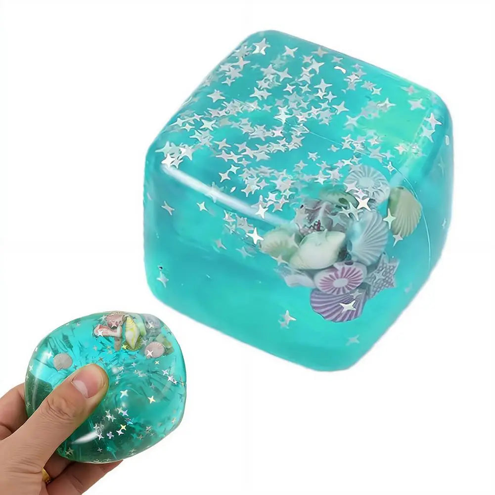 Un cube sensoriel à effet anti-stress en turquoise translucide, de texture apaisante, rempli d’étoiles argentées scintillantes et de coquillages assortis, conçu comme un cadeau attentionné pour soulager le stress.