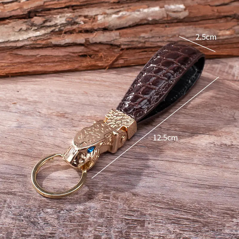 A dark brown crocodile-patterned leather keychain featuring a gold-toned tiger head charm with blue eyes designed as a luxury car key ring.