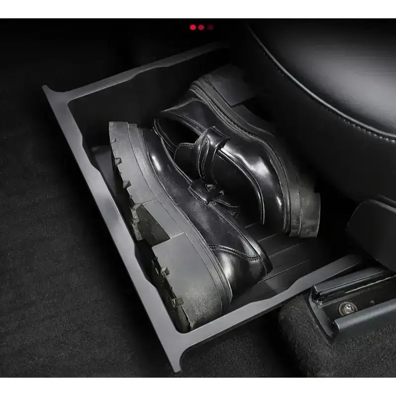 Schwarze Lederschuhe mit kräftiger Sohle, die in einer schwarzen Unter­sitz-Auto-Organizer-Aufbewahrungs-Schubladenwanne in einem Pkw-Fußraum liegen.