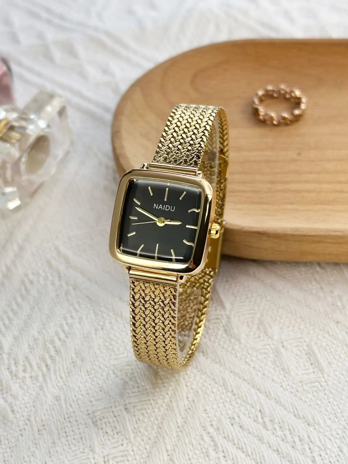 Minimalist square quartz watch for women with a gold mesh bracelet and a black face featuring gold hands and the brand name naidu on its dial.