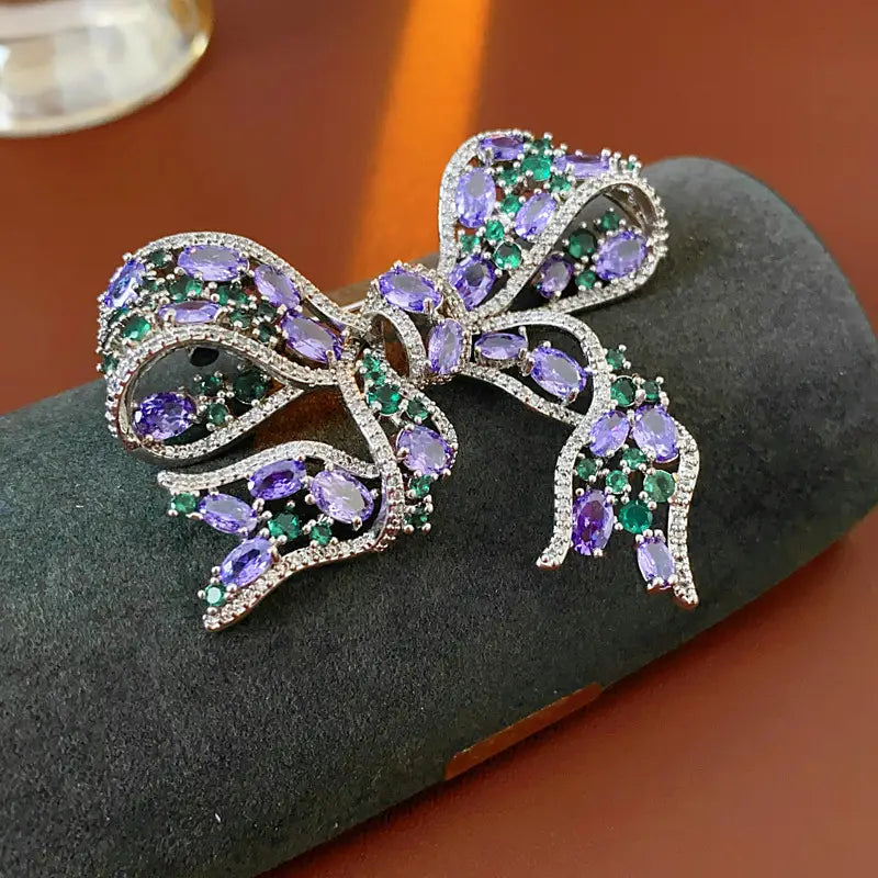 Eine funkelnde silberfarbene Zirkon-Schleifenbrosche, geschmückt mit ovalen lila Amethysten und smaragdgrünen Edelsteinen, die luxuriöses französisches Schmuckdesign ausstrahlt.