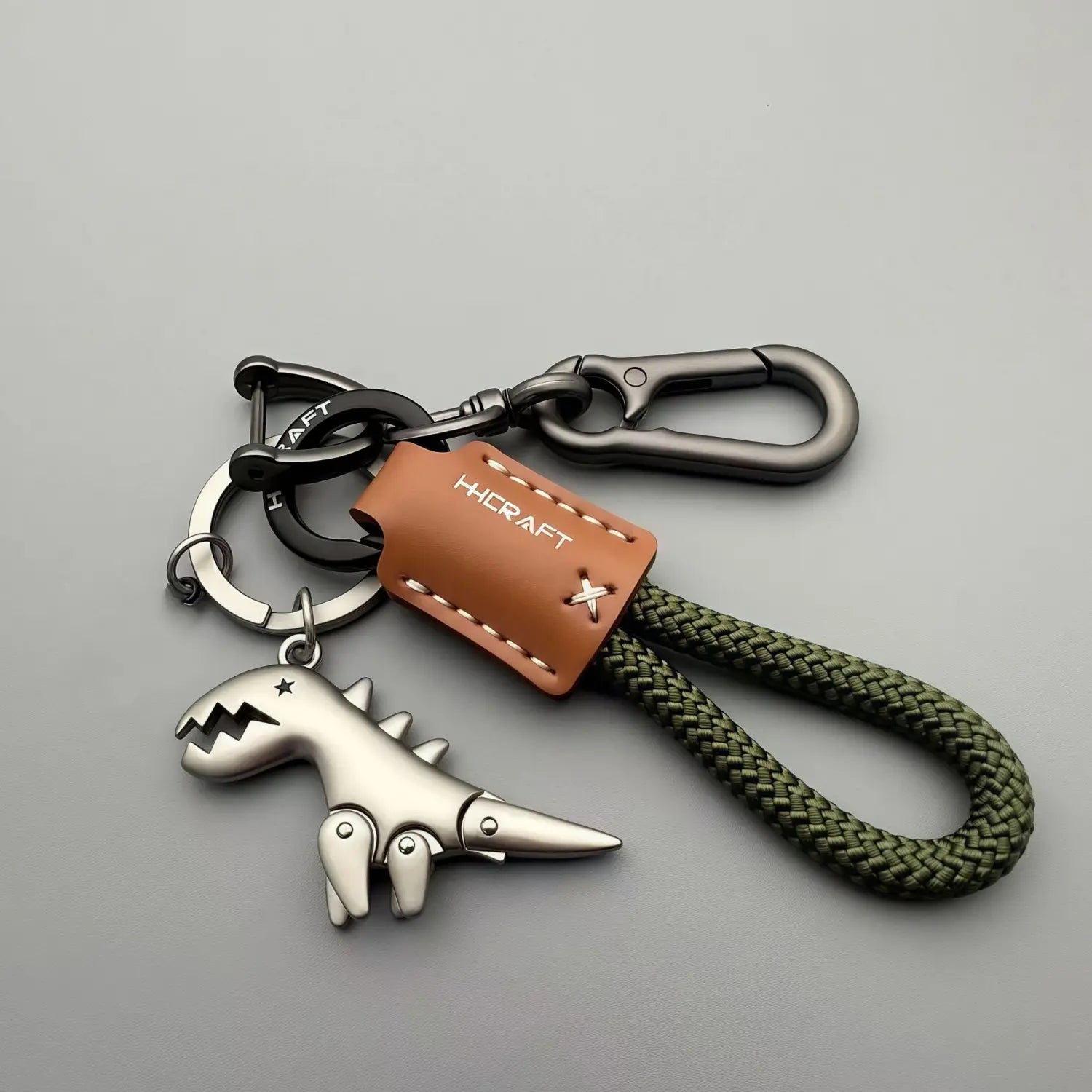 Silver metallic dinosaur keychain with a brown leather tag and olive green braided cord used as a bag pendant.