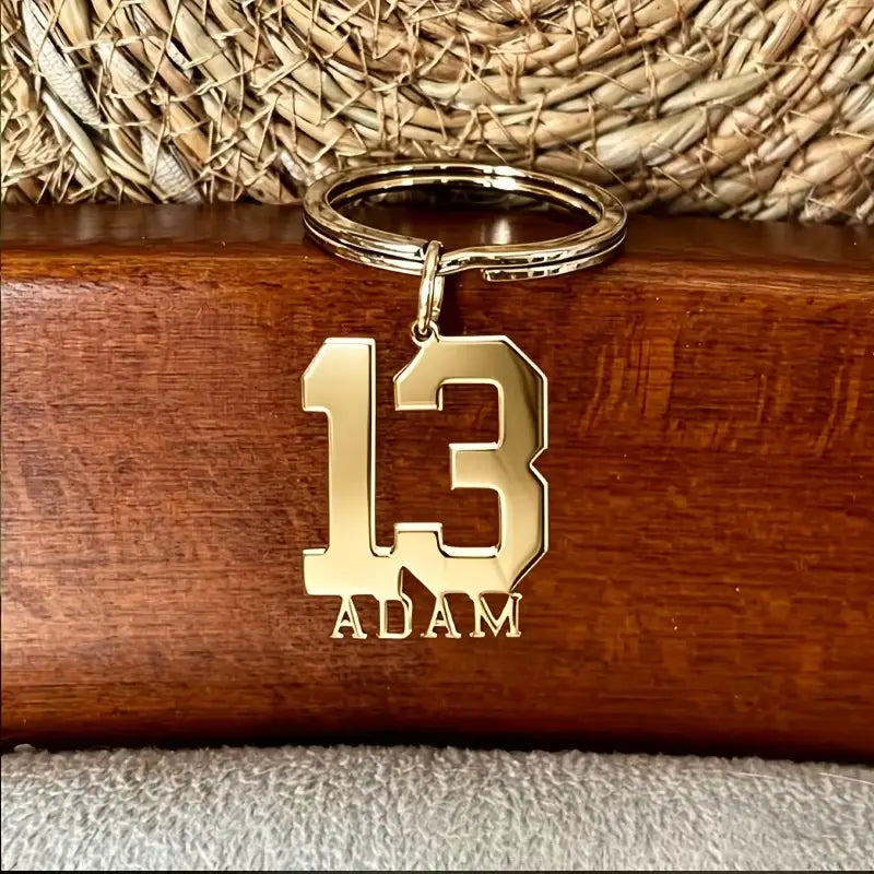 Gold-colored metal custom number and name keychain featuring the number 13 with the name adam engraved below it.