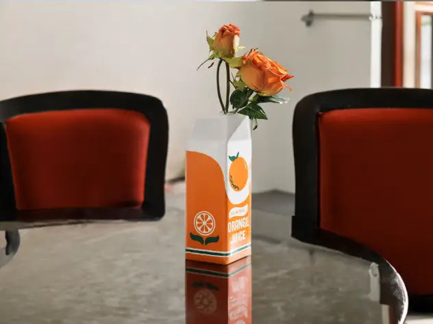 Un vase en forme de contenant rétro en céramique, vibrant orange et blanc, avec une étiquette de jus d’orange rétro, renfermant deux roses orange en pleine floraison.