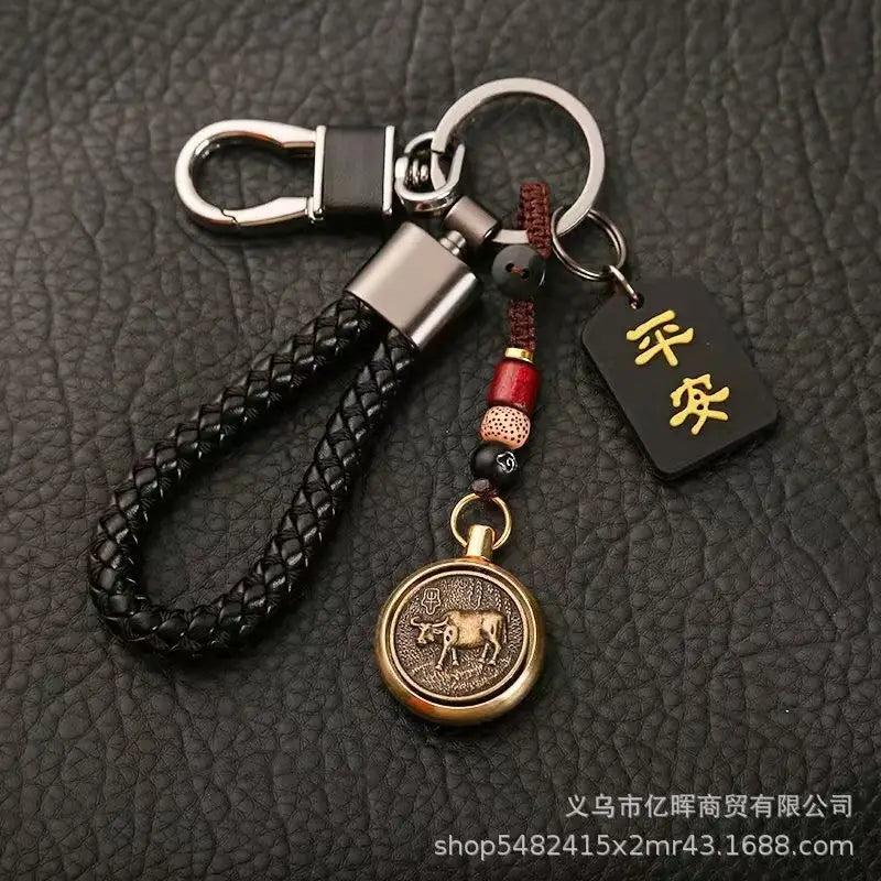 赤と黒のビーズでアクセントを施し、ブロンズ色の円形「牛」ペンダントを備えた黒の編みレザー製キーチェーン。金色の漢字が入った小さな黒いタグ付き（中国の干支をモチーフにした編みレザー製ラッキーペンダントのデザイン）。