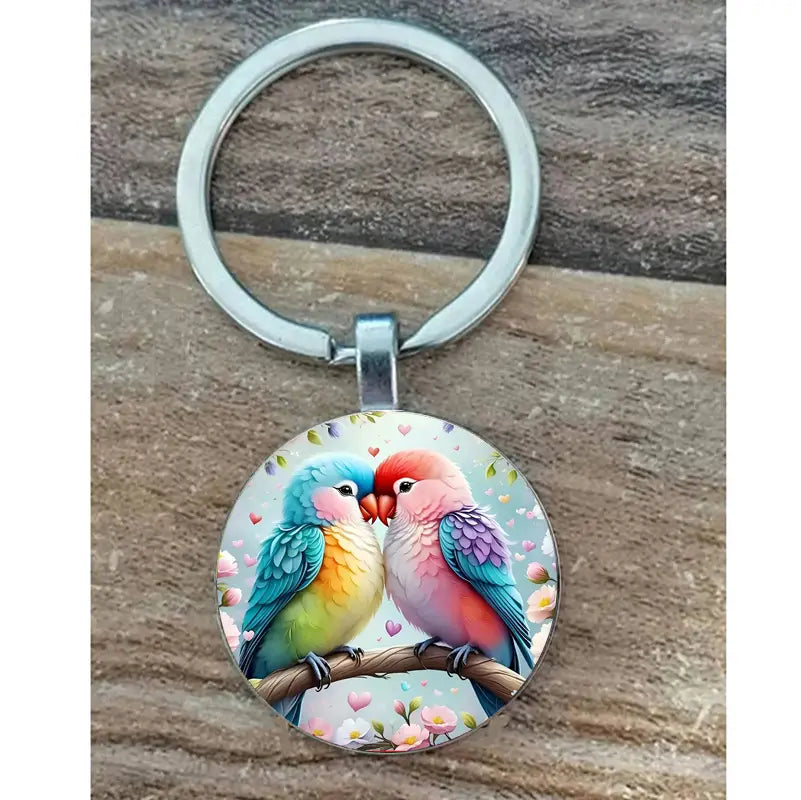 A lovebird keychain glass dome bird keyring featuring a circular clear glass dome with a vibrant illustration of two colorful parrots kissing on a branch attached to a silver metal ring.