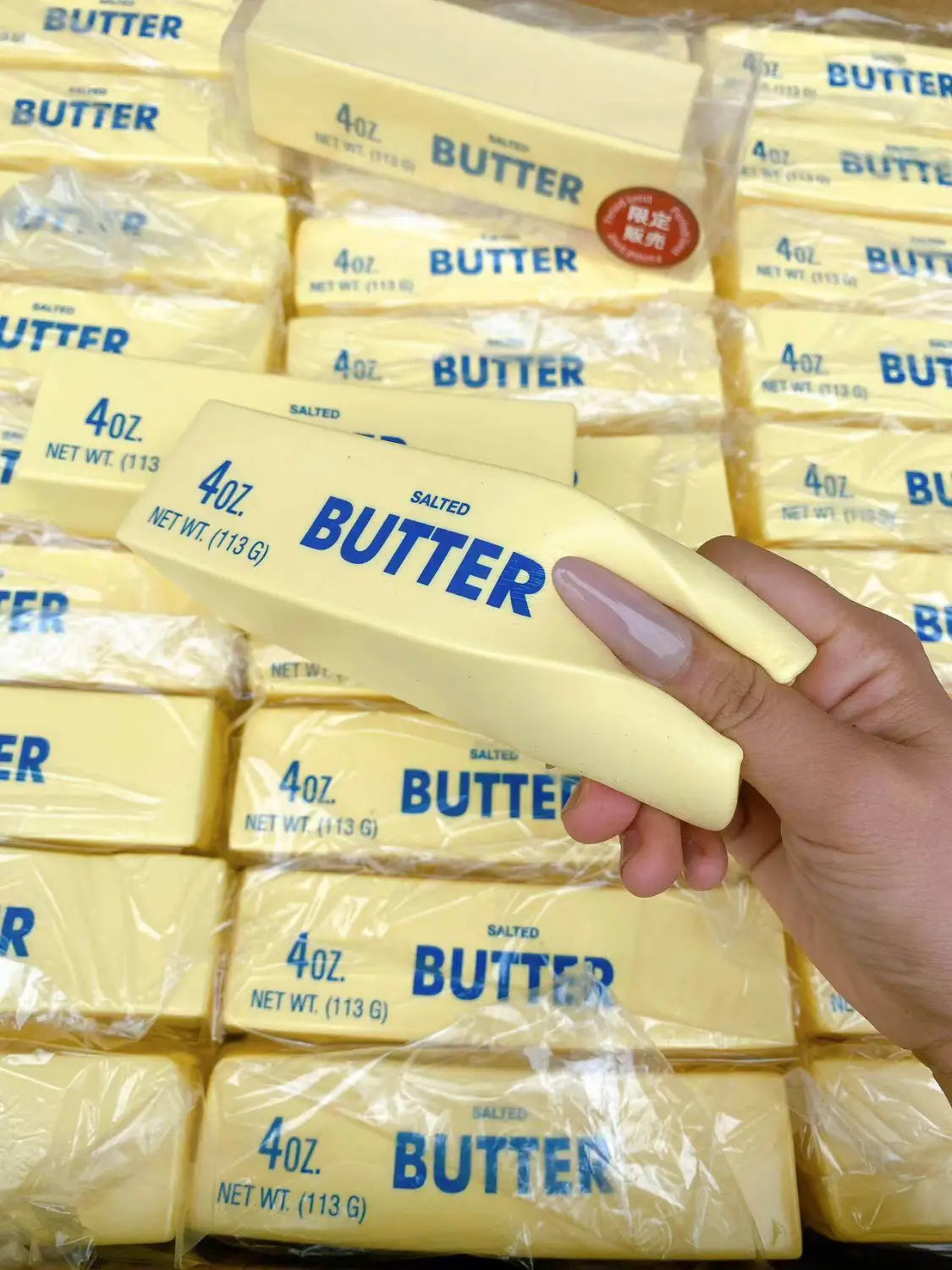 Una mano sostiene un juguete antiestrés blando con forma de barra rectangular de mantequilla que asciende lentamente, de color amarillo pálido, con letras azules y rotulación de 4 oz.