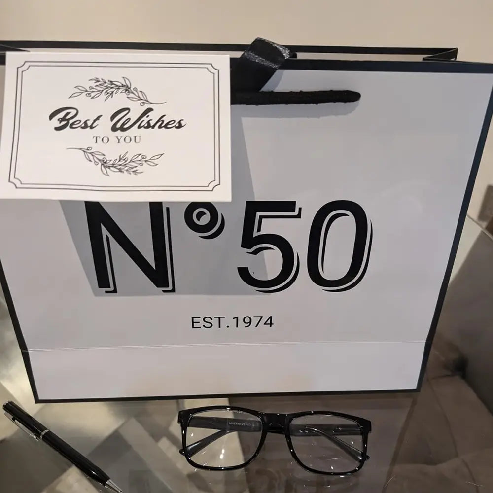 Rechteckige, schwarz gerahmte Brillengläser mit eleganter moderner Gestaltung, dargestellt als Geschenkbeutel zum runden Geburtstag mit Karte und Schleife.