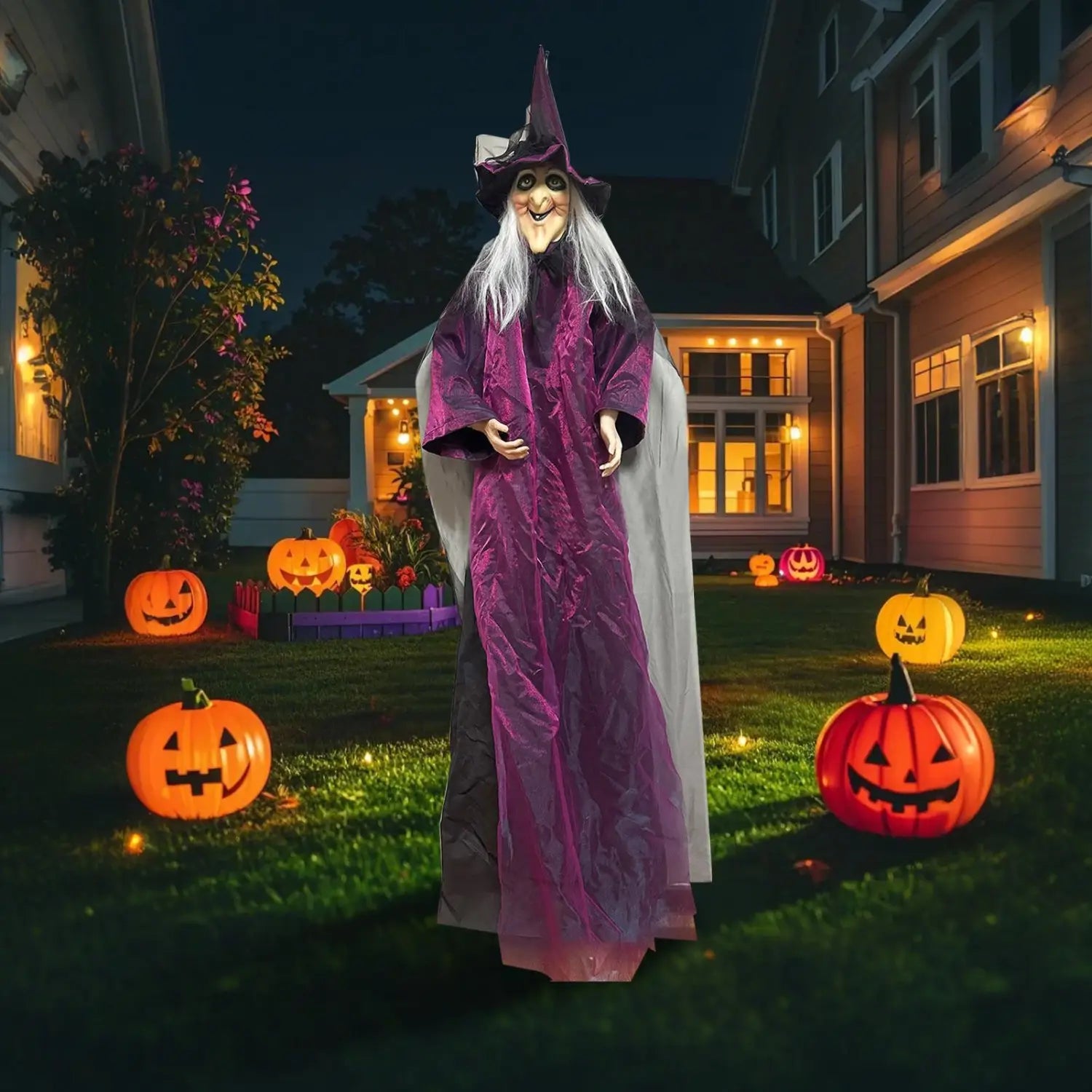accessoire de sorcière d’Halloween à activation sonore grand format, violet et gris, avec un chapeau pointu et de longs cheveux blancs, placé devant une maison entourée de citrouilles jack-o’-lanterns lumineuses