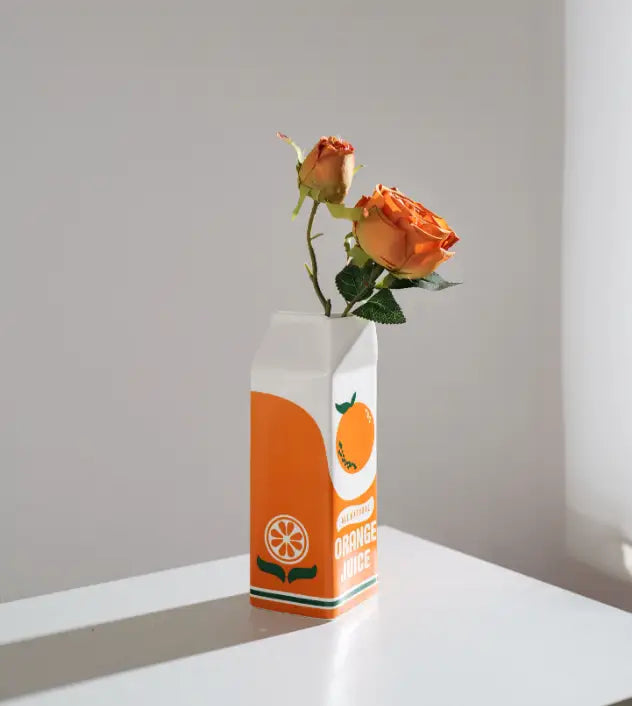 Vase en céramique représentant une brique de jus d’orange, avec un dessus blanc et une bordure verte, arborant un motif d’agrumes orange et une inscription en lettres noires en gras « orange juice ».